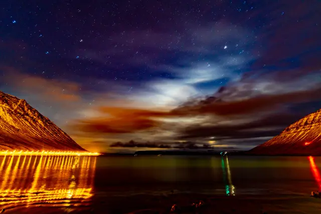 Town lights and stars reflected in a calm fjord framed by dark mountains, Iceland