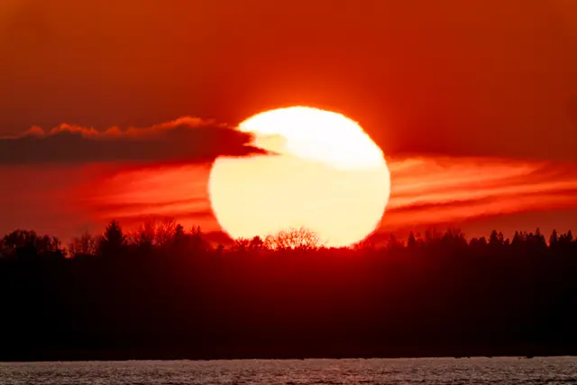 A vivid red sunset sinking behind a silhouetted shore.