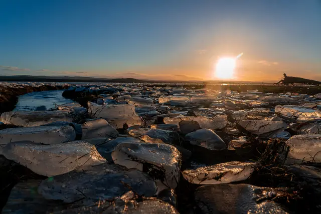 A boulder-strewn shoreline under a bright dawn sky.